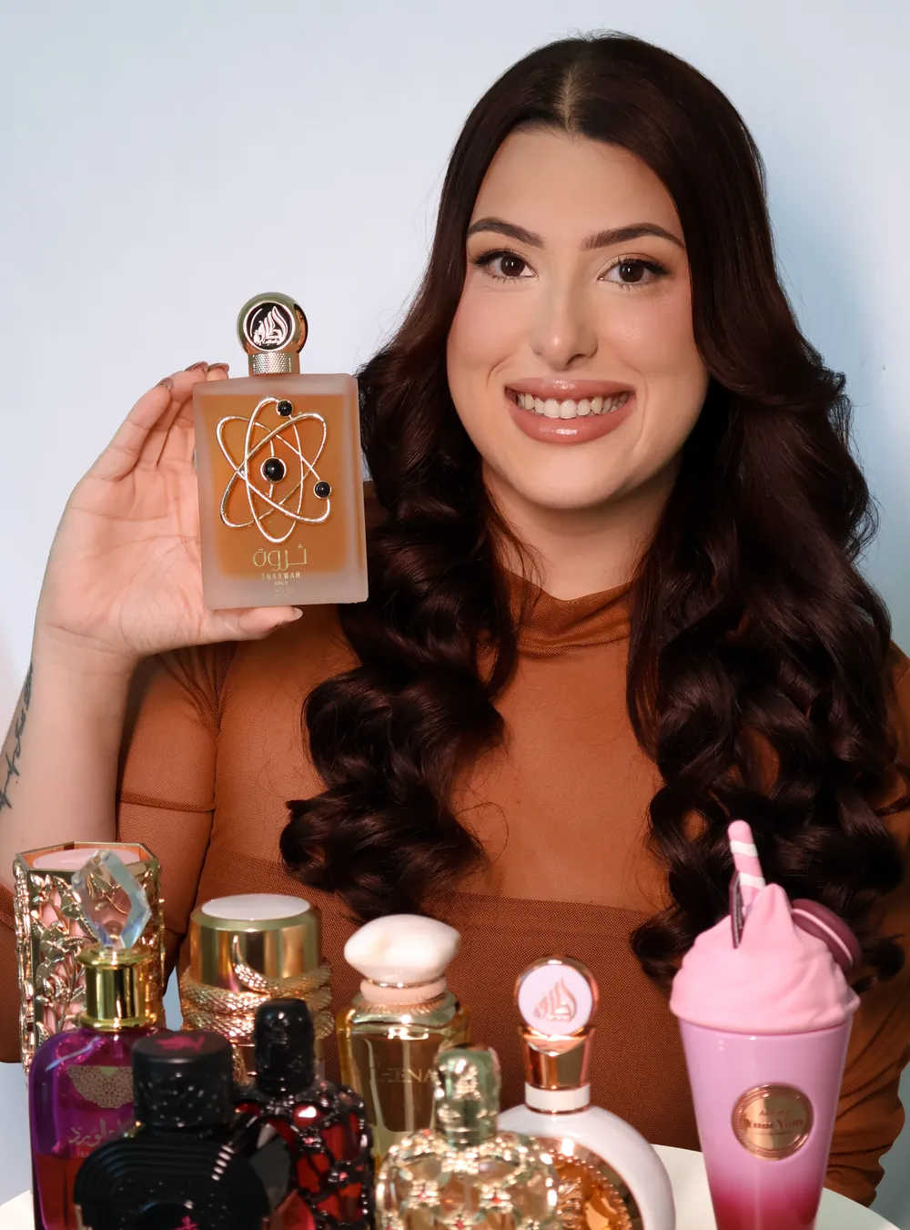 Aninha Barros sorrindo, segurando um perfume, com vários perfumes árabes sobre a mesa
